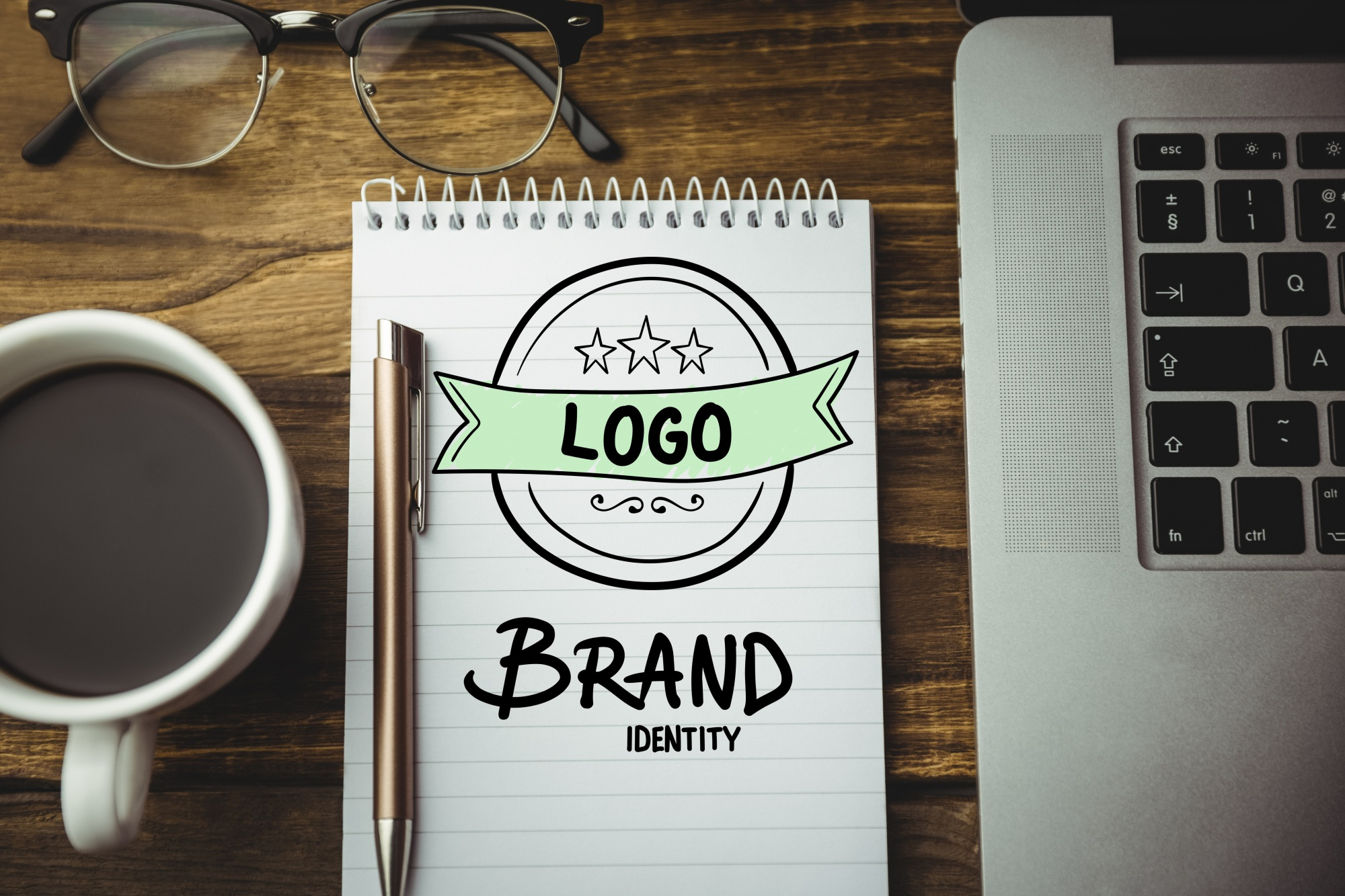 Bloco de notas com "LOGO" e "BRAND IDENTITY" escritos, simbolizando o planejamento de marca, criação de identidade visual e estratégia de branding, ao lado de óculos, caneta, xícara de café e laptop.