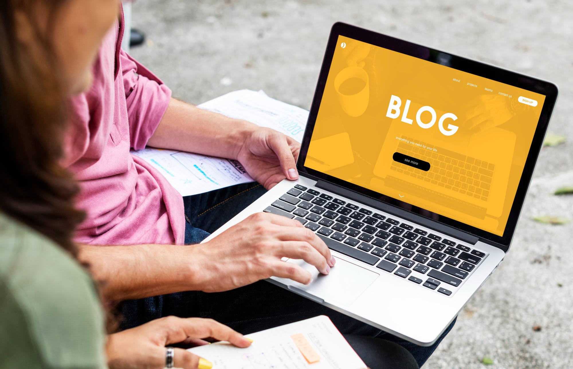 Duas pessoas colaborando em um laptop que exibe a página "BLOG", simbolizando a criação de conteúdo, escrita e estratégia de marketing digital.